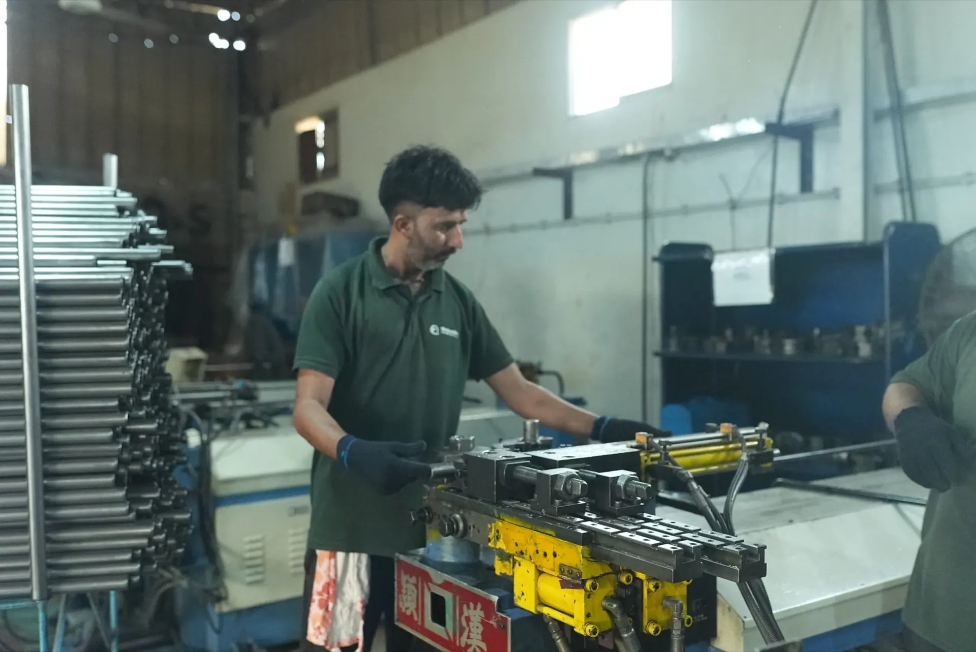 Operator at a hydraulic pipe bender beside a rack of straight tubes